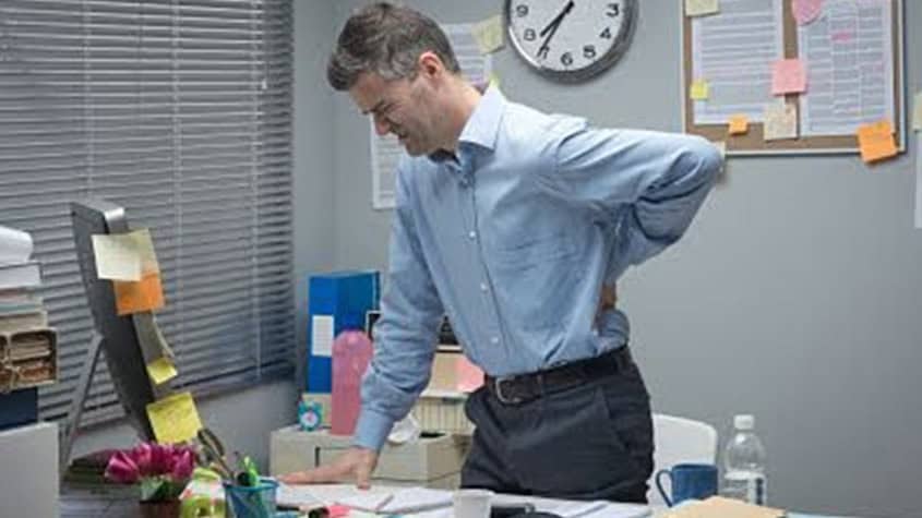 Man standing at his desk with a pained expression, gripping his lower back