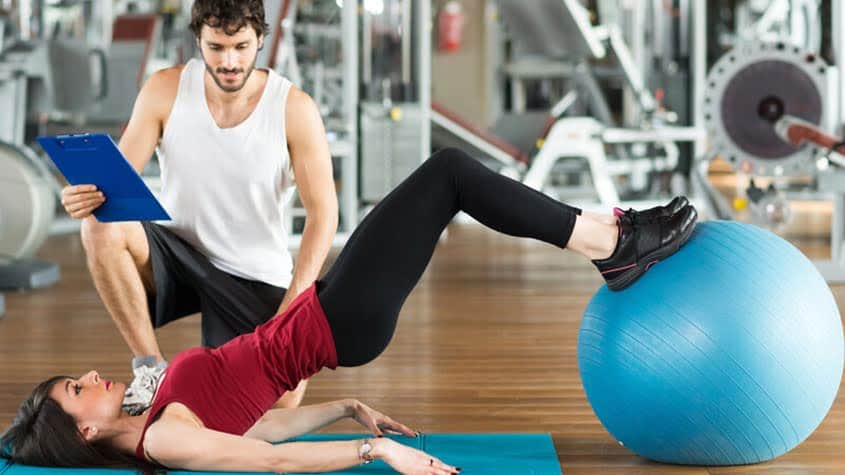 Woman doing core-strengthening exercise on a blue stability ball with trainer assistance.